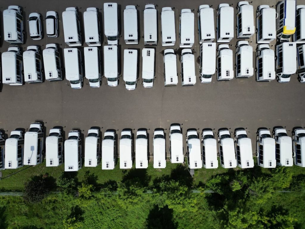 Home Aerial view of a large parking lot filled with white vans on a sunny day.